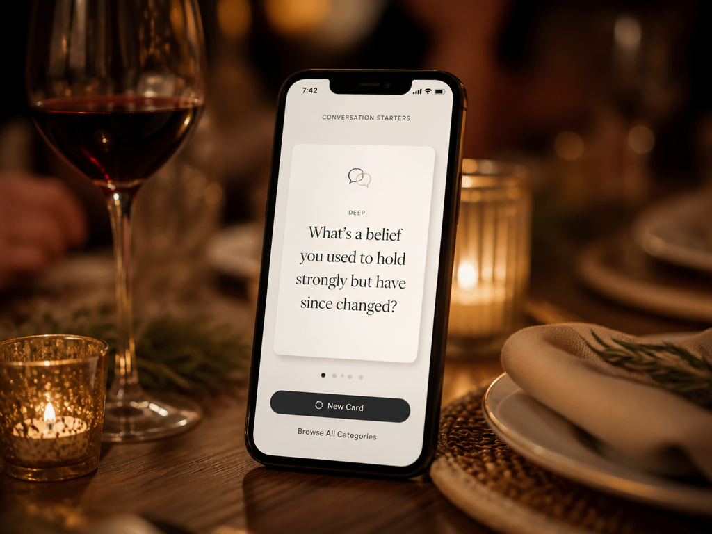 A smartphone displaying a digital conversation starter card resting on a candlelit dinner table between courses