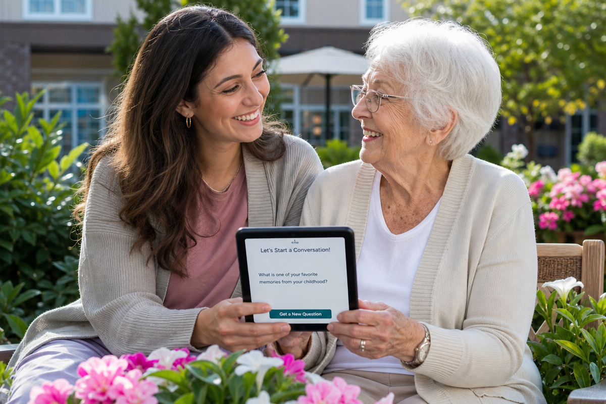 A young adult and an elderly woman sitting together in a garden at a senior living facility, looking at a tablet with a conversation starter app and smiling warmly at each other.